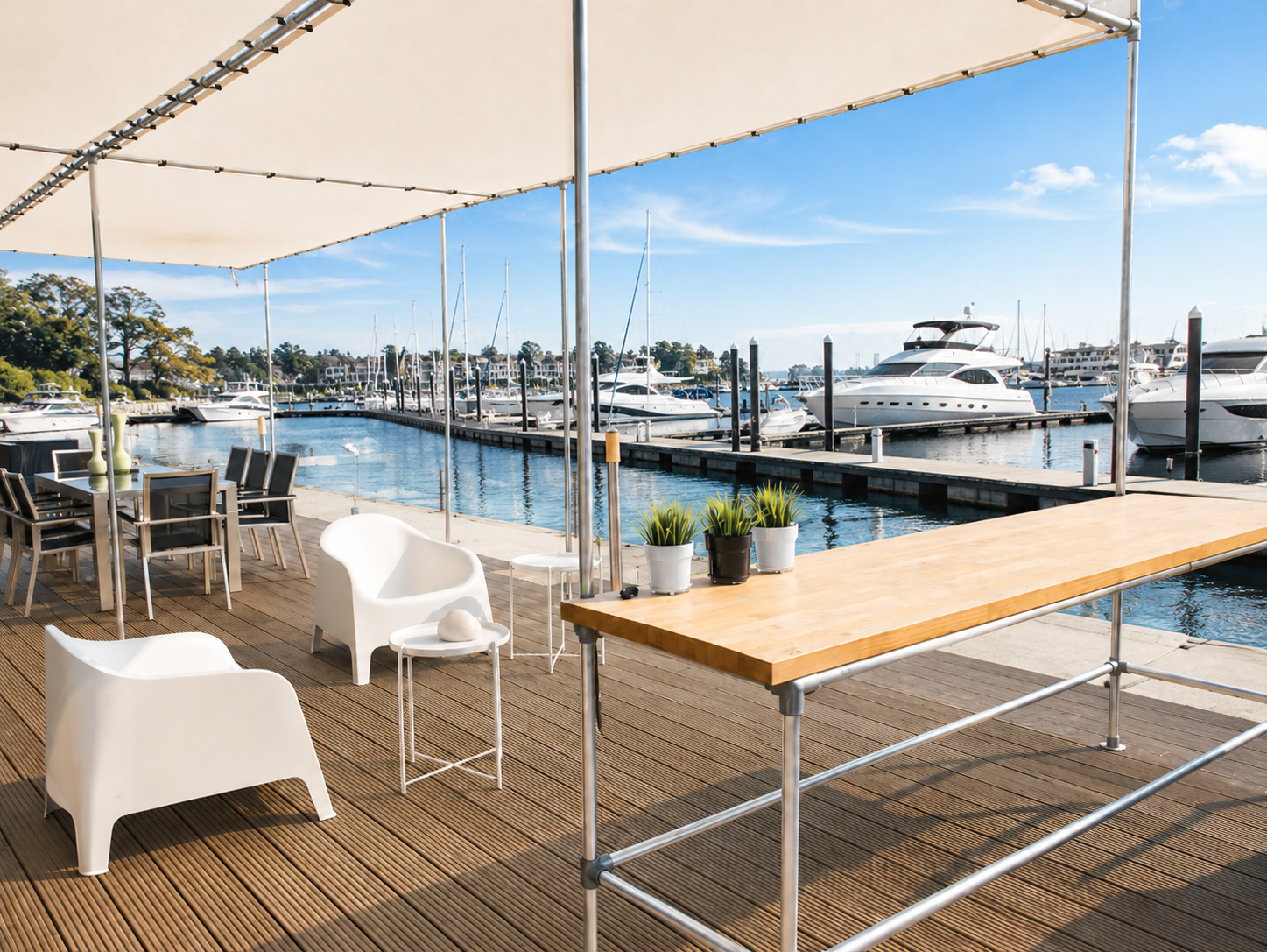 Holzterrasse unter Sonnensegel am Yachthafen. Moderne weiße Lounge-Sessel, Esstisch und langer Holztresen mit Topfpflanzen. Im Hintergrund viele Segelboote und Yachten im blauen Hafenwasser bei strahlend blauem Himmel.