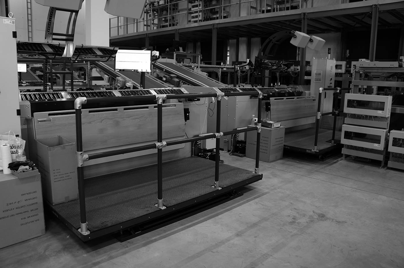 Schwarz-Weiß-Ansicht einer industriellen Sortieranlage mit mehreren Förderbändern, die von links nach rechts in den Hintergrund verlaufen. Im Vordergrund eine erhöhte Arbeitsplattform mit schwarzen Handläufen. Rechts gestapelte Steuergeräte.