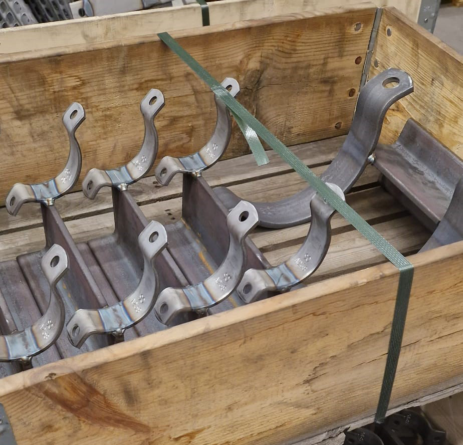 Mehrere identische, gebogene Metallhalterungen mit angeschweißten Befestigungslaschen liegen in einer hölzernen Transportkiste. Die grauen Teile zeigen leichte Schweißverfärbungen und eingelaserte Kennzeichen. Grüne Umreifung sichert den Inhalt.