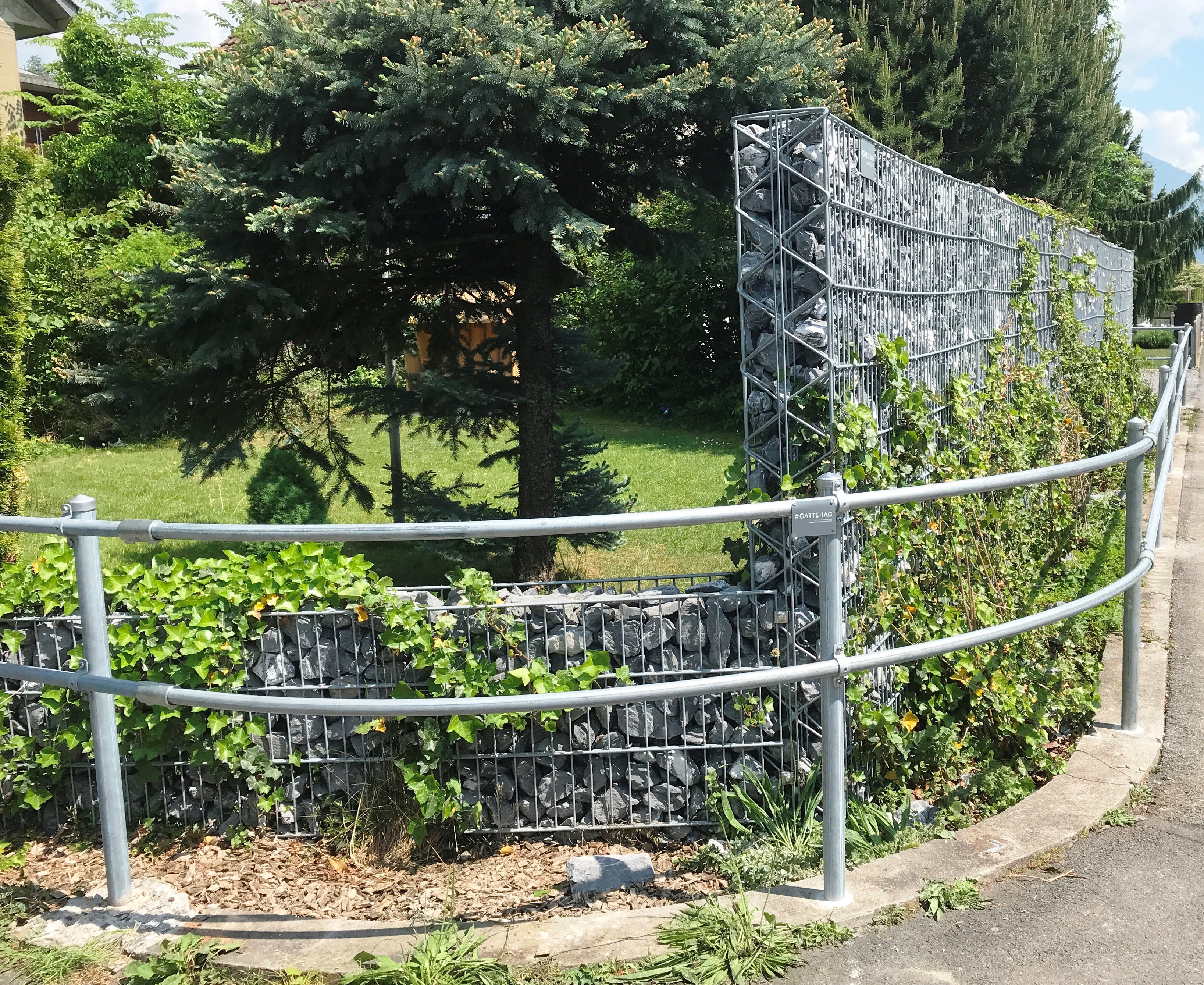 Eine Gabionenwand aus Drahtgeflecht und grauen Steinen trennt einen gepflegten Garten von einem Gehweg. Sie ist teils mit üppigem grünem Efeu bewachsen. Davor verläuft ein geschwungener, silberner Metallhandlauf.