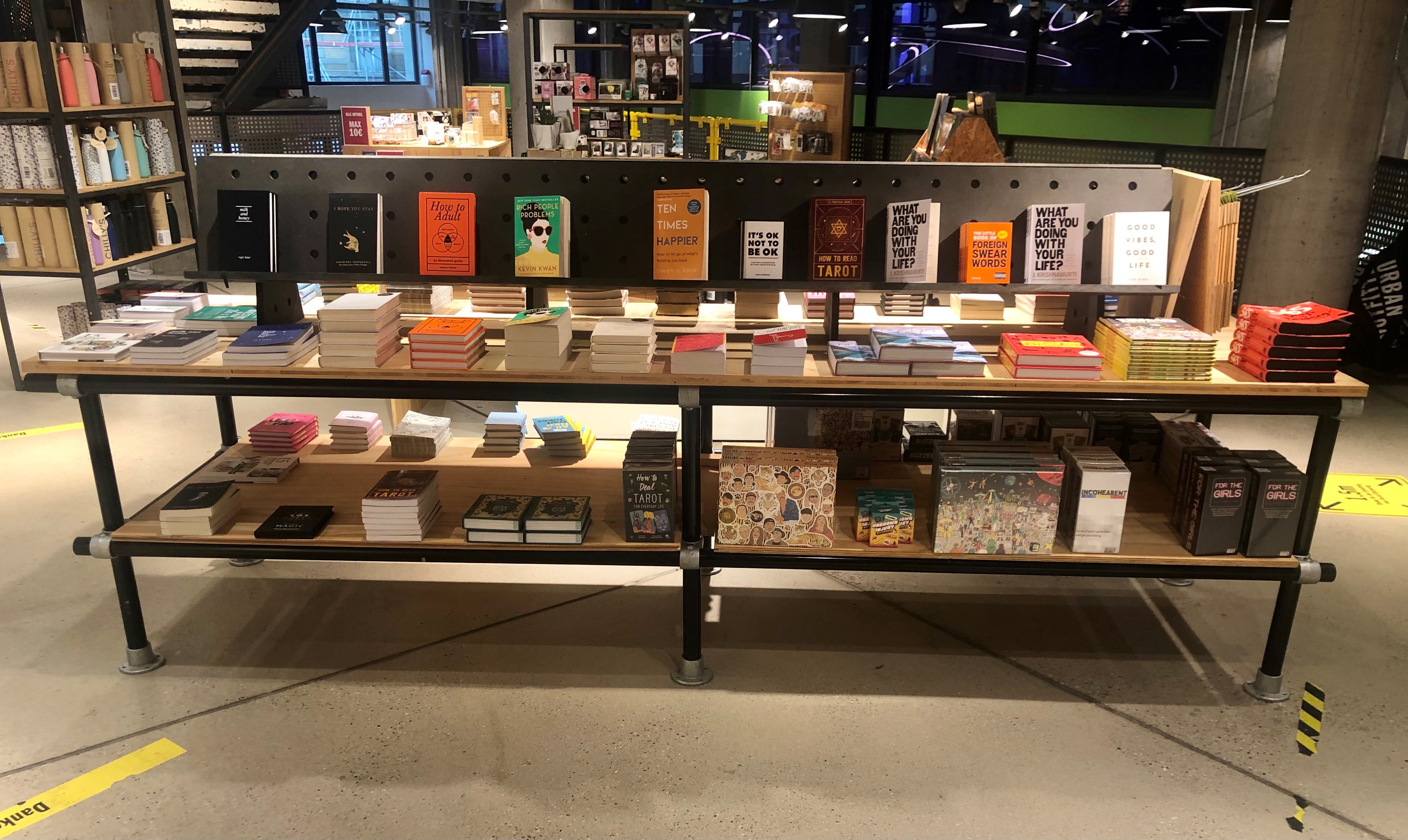 Großer moderner Verkaufsstand im Geschäft mit Büchern und Geschenkartikeln. Oben viele Bücher mit Cover nach vorn, u.a. Selbsthilfe- und Lifestyle-Bestseller. Darunter weitere Bücherstapel und dekorative Boxen mit Spielen oder Karten.