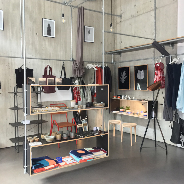 Stilvolles Geschäft im Industrial-Design: rohe Betonwände, Metallrohr-Regale. Taschen, Kleidung, Keramik, gefaltete Textilien auf Holzregalen. Kunstdrucke an den Wänden; Glühbirnen von der Decke. Rechts Teleskop und Holzschemel.