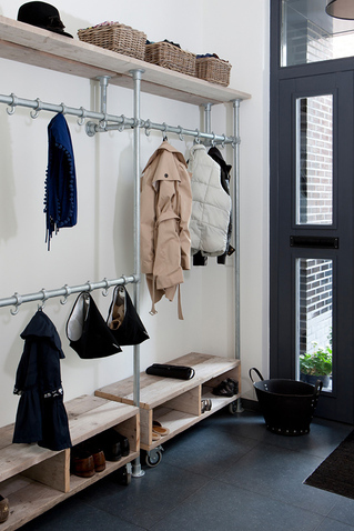 Heller Flur mit Garderobe im Industriestil: Metallrohre, Holzablagen. Oben Körbe und Hut. Hängend blauer Überwurf, beiger Trenchcoat, weiße Weste. Unten Schuhe. Rechts dunkle Glastür.