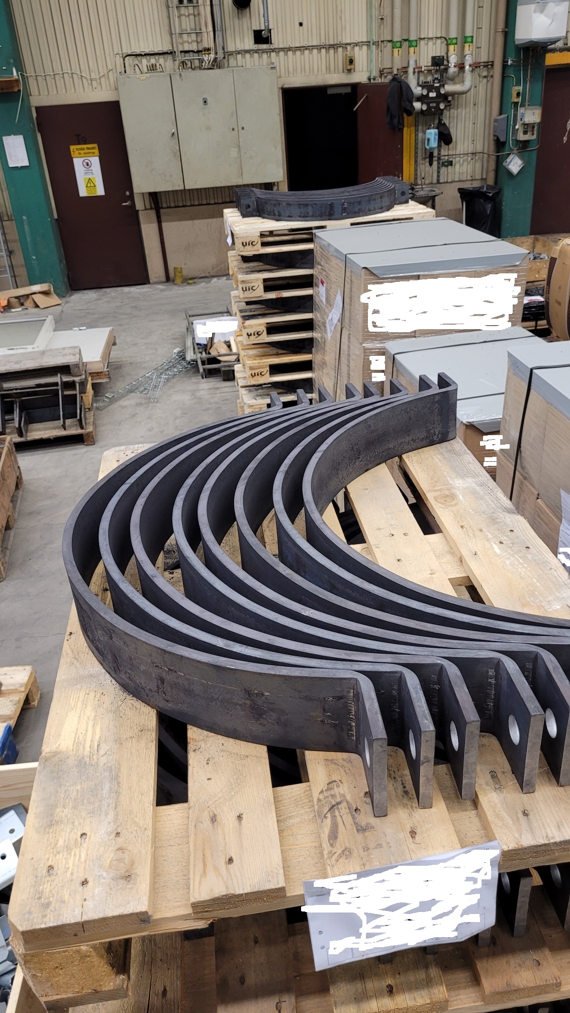 Dunkelgraue, schwere, gebogene Metallteile liegen auf einer Holzpalette in einer Industriehalle. Dicke C-Bügel sind geschwungen angeordnet, mit flachen Enden und Befestigungslöchern. Im Hintergrund Paletten und Industrieausstattung.