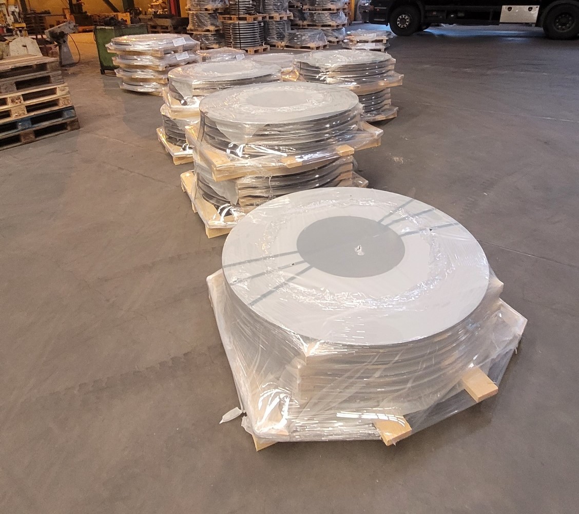 Große, runde Metallplatten, gestapelt und in Folie verpackt, stehen auf Holzpaletten in einer Lagerhalle. Die Stapel sind in mehreren Reihen angeordnet. Im Hintergrund ist ein LKW zu erkennen, links liegen leere Paletten.