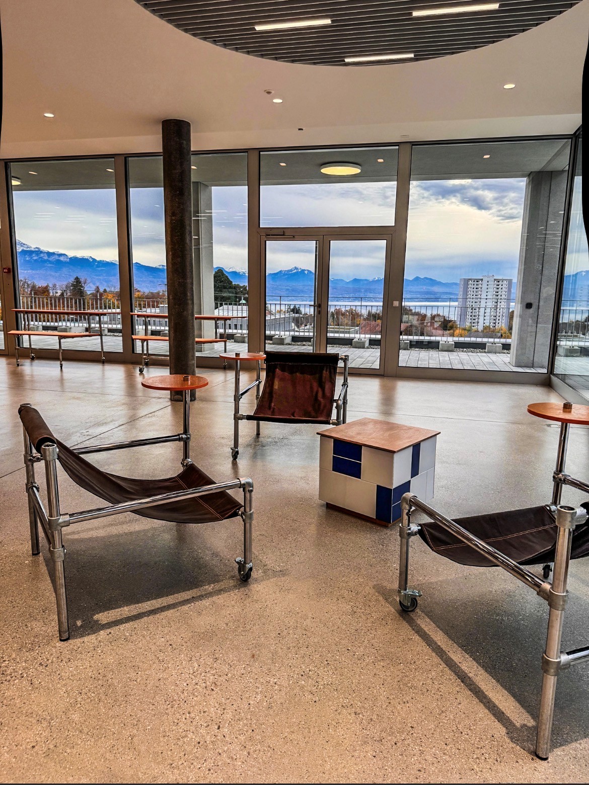 Moderner Lounge-Bereich mit großen Fenstern und weitem Blick auf See, schneebedeckte Berge und bewölkten Himmel. Drei braune Ledersessel mit Chromgestell auf Rollen sowie kleine orange Tische stehen auf hellem Terrazzoboden.
