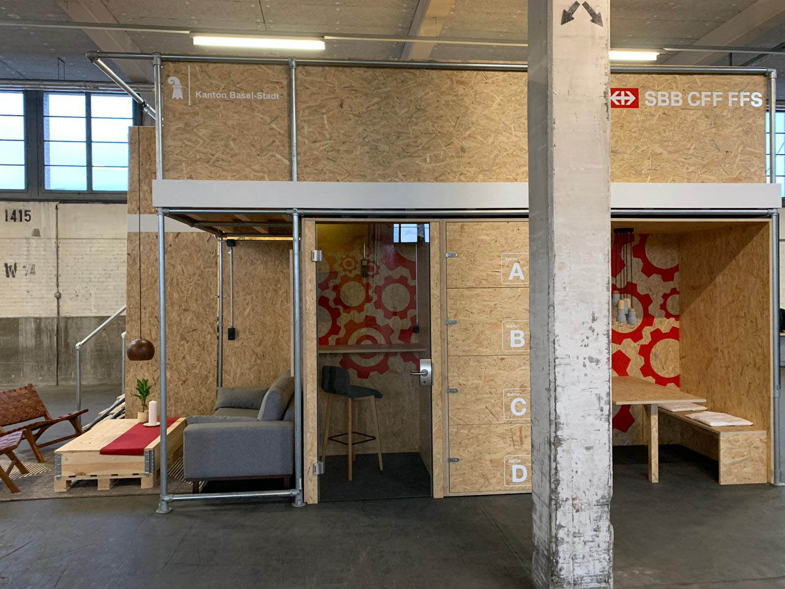 Modularer Arbeits- und Lounge-Bereich aus OSB-Platten und Metallgerüst in großer Halle. Links Sofa mit Palettentisch. Mittig Kabine mit Bartheke und Schließfächern. Rechts Kabine mit Holztisch und Bank. Logos SBB und BS sichtbar.