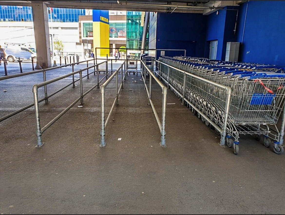 Zugangsbereich eines IKEA-Geschäfts: rechts eine lange Reihe sauber gestapelter Einkaufswagen mit blauen Griffen. Links führen metallene Absperrungen über den grauen Boden zum Eingang. Im Hintergrund großer gelb-blauer IKEA-Schriftzug.
