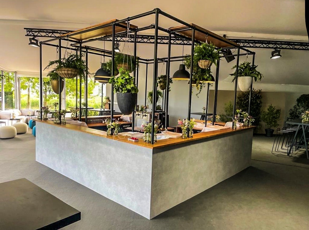 Moderner Tresen mit grauer Betonoptik und Holzplatte in hellem Raum. Darüber schwarze Metallkonstruktion mit vielen hängenden Grünpflanzen und Pendelleuchten. Tresen mit kleinen Blumenarrangements. Große Fenster mit grüner Landschaft.