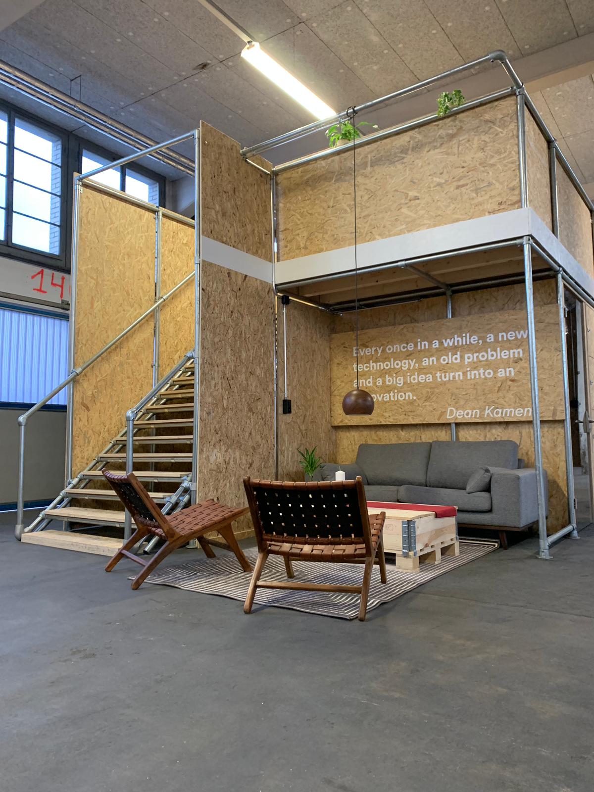 Kreativer Innenraum mit zweistöckiger Holzkonstruktion auf Metallrohren. Unten gemütliche Sitzecke mit grauem Sofa, zwei braunen Lederstühlen und Paletten-Couchtisch. An der Wand ein Innovationszitat. Links Holztreppe nach oben, Pflanzen.
