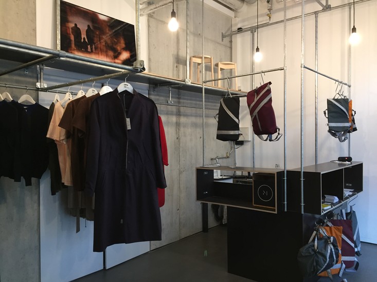 Modernes Bekleidungsgeschäft: Links Kleidung und Kunst. An Rohren hängen Rucksäcke mit Reflexstreifen. Schwarzer Tresen mit Logo. Industrielles Licht durch Glühbirnen.