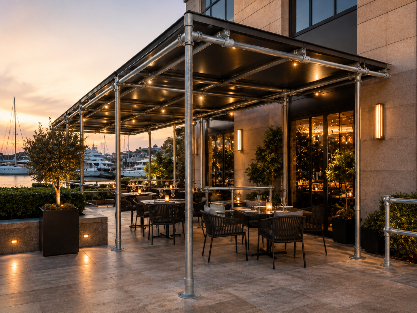 Restaurantterrasse am Yachthafen bei Sonnenuntergang. Überdachung mit Lichterketten, Tische mit Gläsern und Kerzen. Boote im Hintergrund, orange-violetter Himmel, Pflanzen und Bodenlichter.