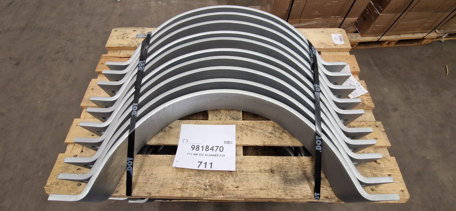 Große, gebogene, silberne Metallklammern liegen ordentlich auf einer Holzpalette, mit schwarzen "DOT"-Gurten gesichert. Vorn ein Etikett: "9818470" und "711 MM SSG KLAMMER FZV". Bereit für Transport im Lagerhaus.